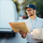 Lachende bezorger met pakket en envelop voor een bestelbus, passend bij flexibel werk als bezorger.