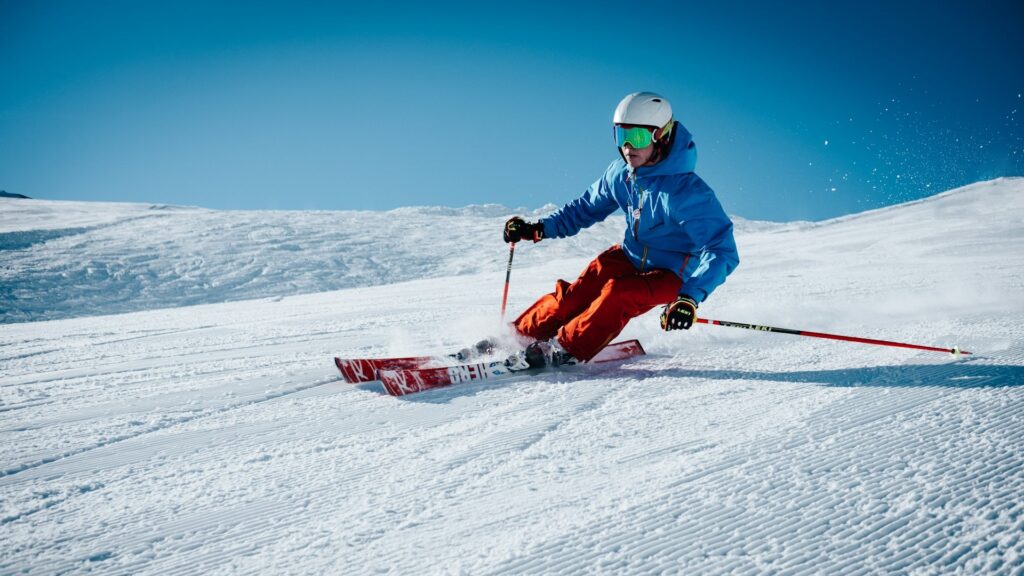 Skiër met complete wintersportuitrusting maakt een bocht op een besneeuwde piste onder een heldere blauwe lucht.