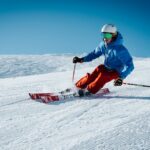 Skiër met complete wintersportuitrusting maakt een bocht op een besneeuwde piste onder een heldere blauwe lucht.