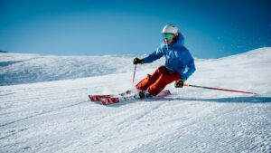 Skiër met complete wintersportuitrusting maakt een bocht op een besneeuwde piste onder een heldere blauwe lucht.
