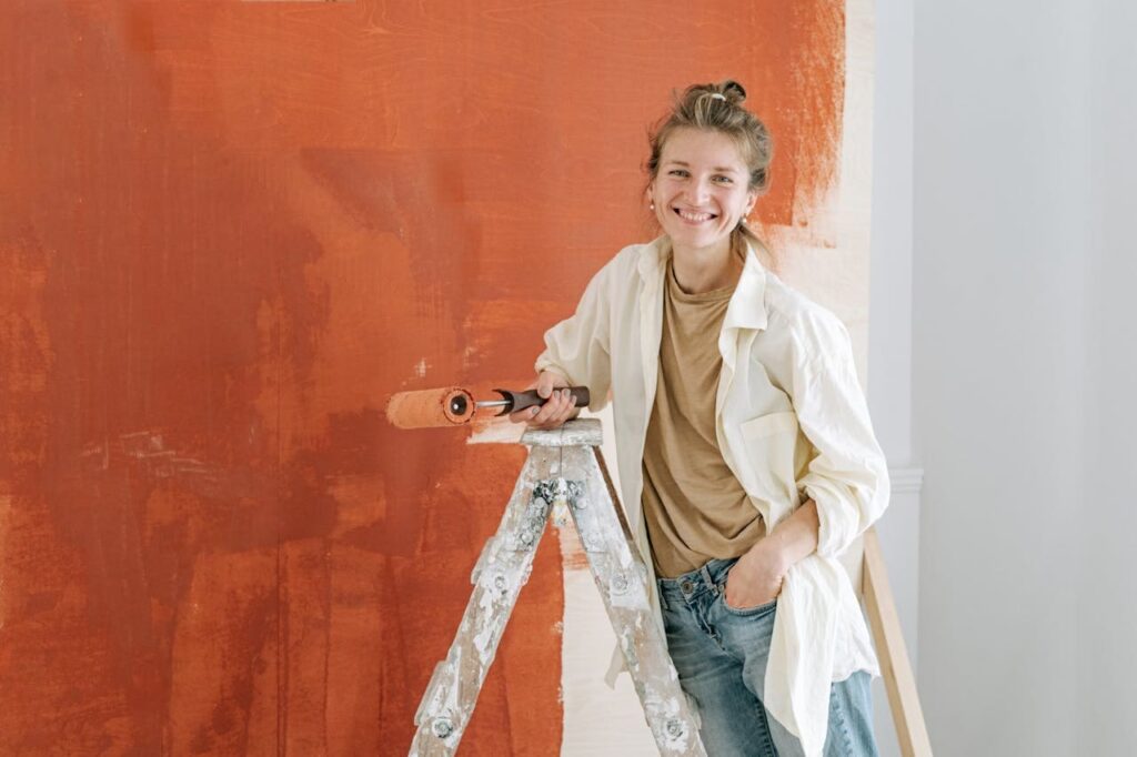 Vrouwelijke binnenschilder staat lachend op een ladder en schildert een muur in een warme terracotta kleur