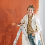 Vrouwelijke binnenschilder staat lachend op een ladder en schildert een muur in een warme terracotta kleur